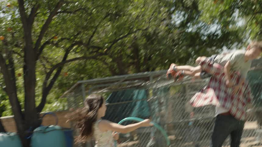 A young girl chasing a man, who has a young boy thrown over his shoulder, with a water hose