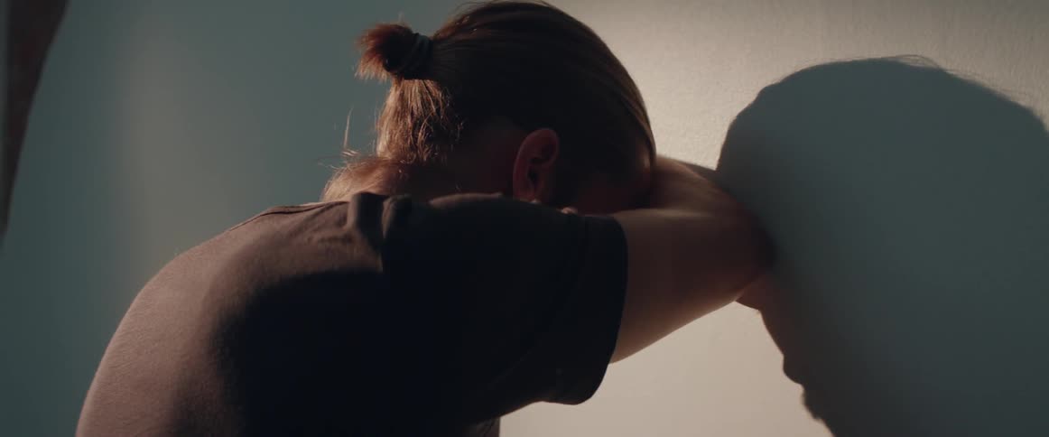An exhausted person leans heavily against the bedroom wall under warm dramatic light.