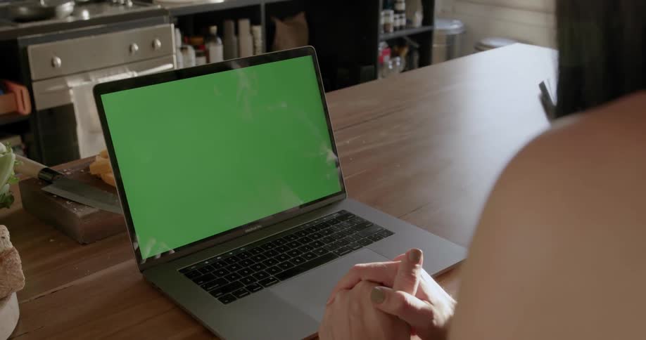 Woman sits looking at a laptop with a green screen and another person types on it