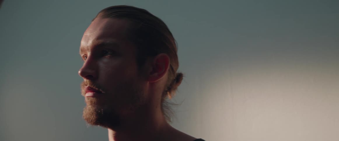 A serious young man looks directly ahead blankly under soft atmospheric indoor lighting.