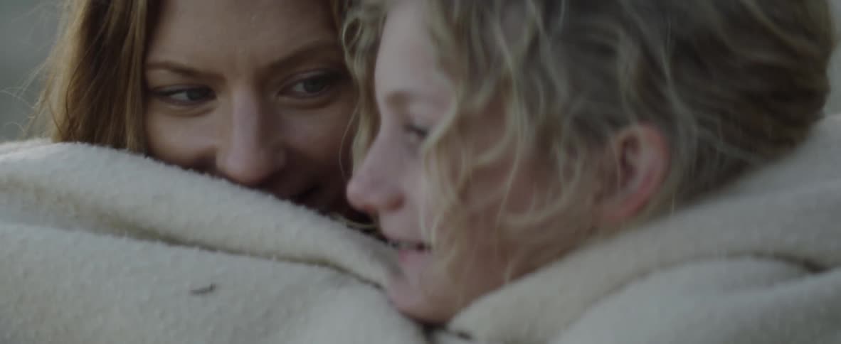 A mother and daughter are laughing and snuggling up under a blanket outside