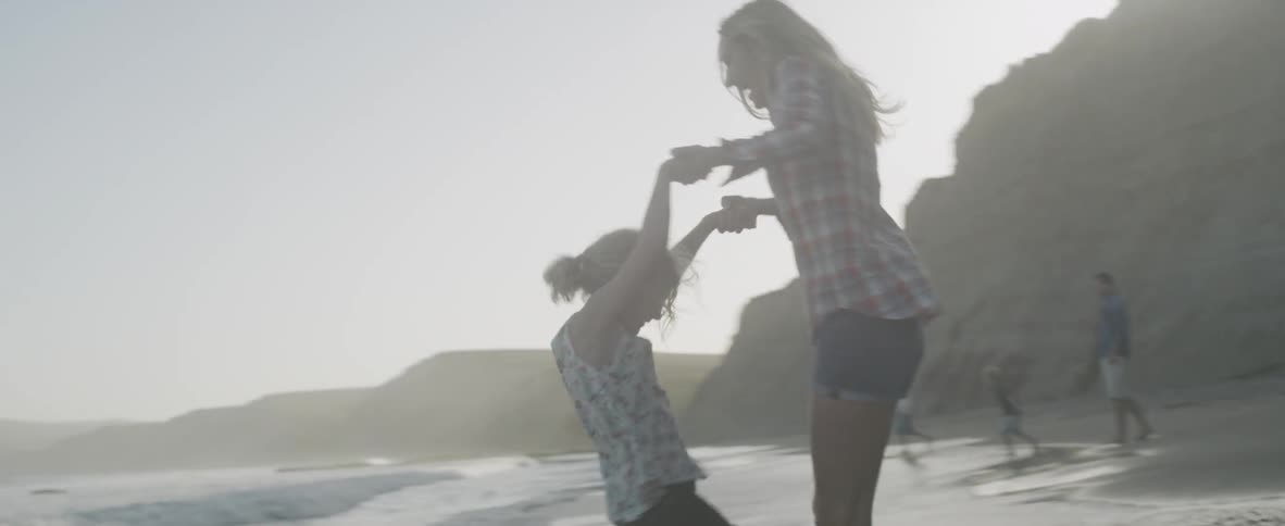 A mother and daughter spin and laugh together along the shore at sunset