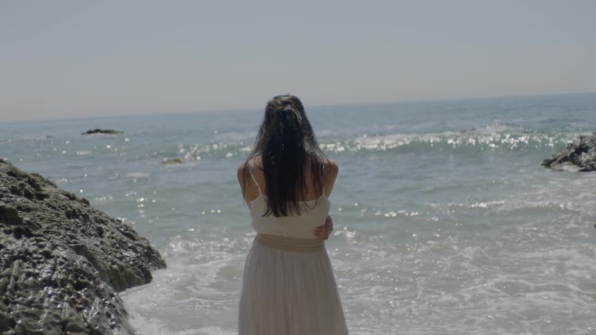 A woman in a white dress stands alone on the rocky beach staring out into the ocean