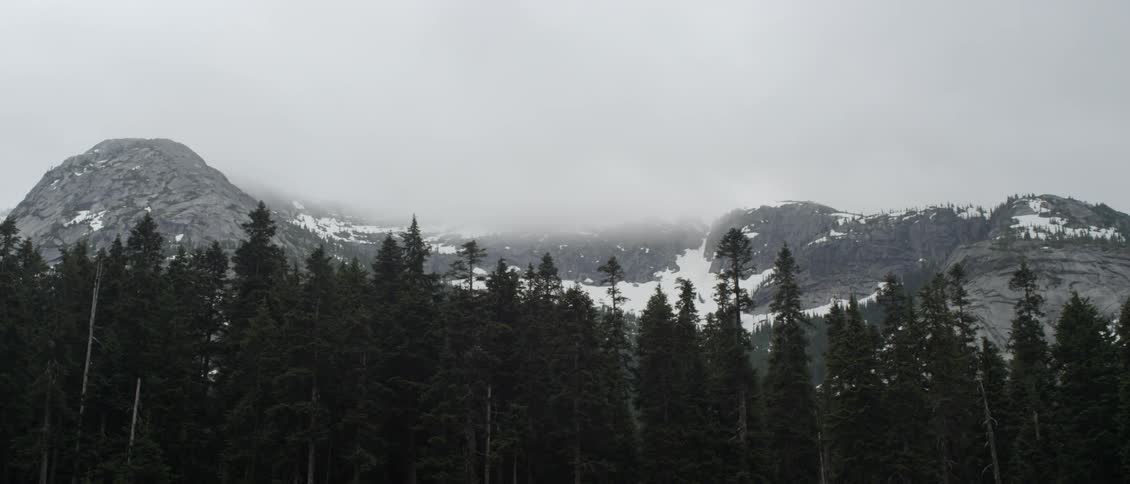 Clouds touch the tops of snowy mountains with trees in the front