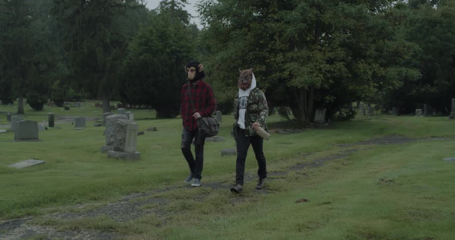 Two people wearing animal masks are walking through cemetery
