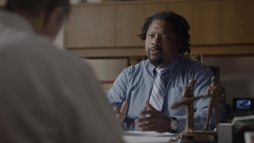Man wearing a striped tie sits at a desk talking to another man