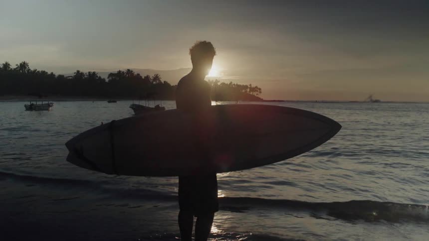 A surfer is standing at the water's edge with his surfboard at sunset
