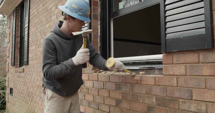 Man wearing a hard hat uses a chisel to tap on bricks on the outside of a house