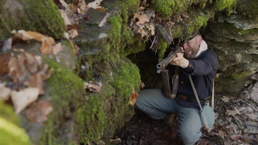 Soldier hides under a mossy clump of rocks aiming his rifle
