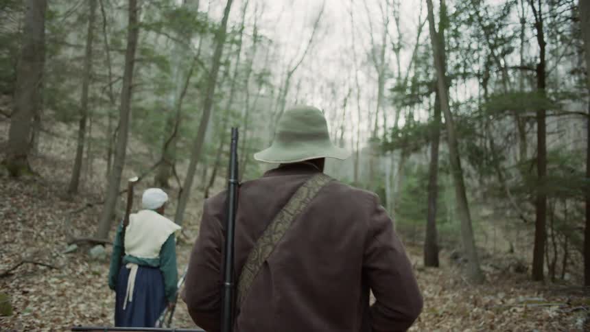 A man and woman carry rifles through the woods