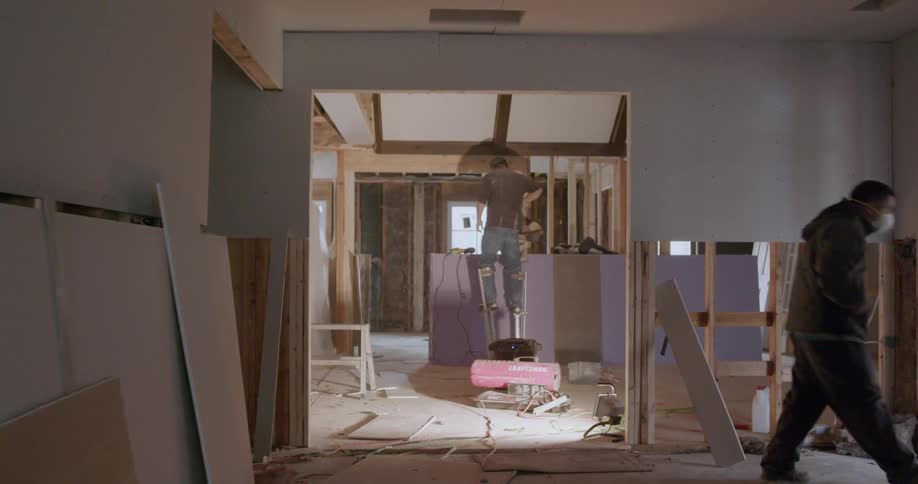 Man wearing a mask walks through a house he is renovating as another man stands on stilts