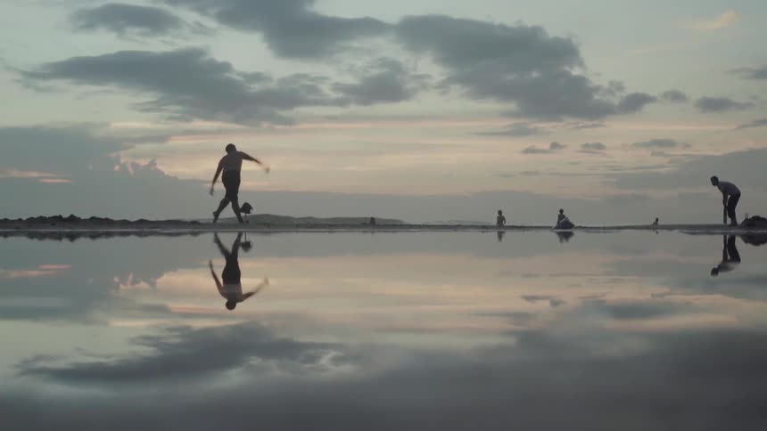 A bowler winds up the ball and throws it towards a batter on the beach