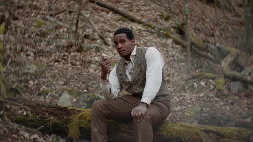 A man sits on a fallen tree and smokes a pipe