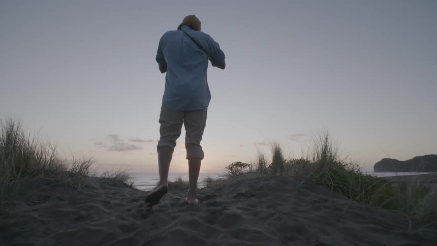 A man climbing to the top of a sand dune and photographing the sun setting over the ocean