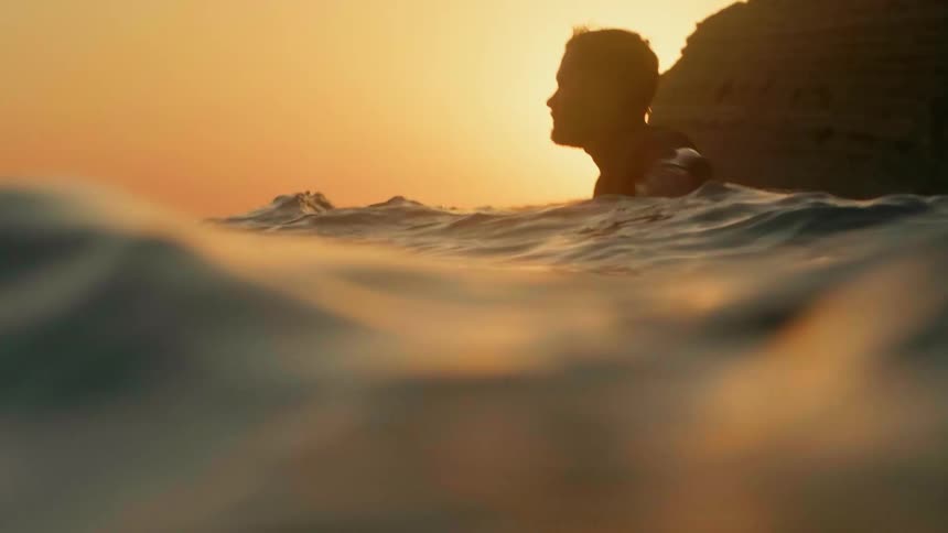 A surfer is paddling out through the waves at sunset