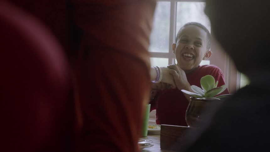 A boy is laughing and playing as someone tries to feed him at a table in daylight