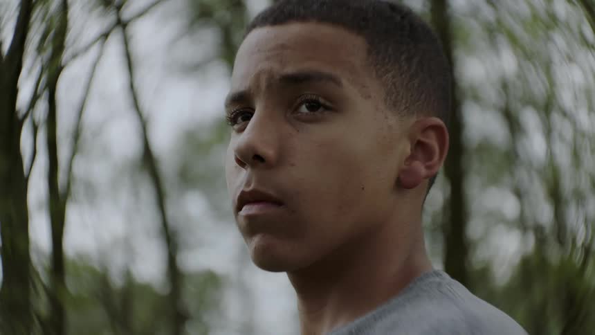A boy stands outside with trees behind him