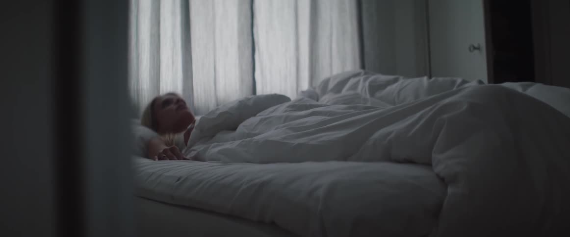 An awake blonde woman lies restless under white duvet on bed.