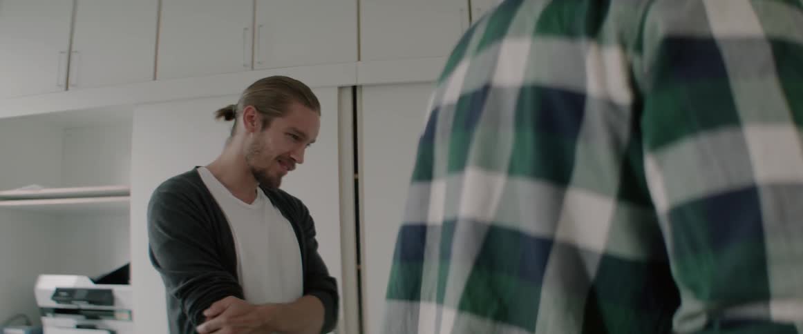A smiling young man looks down and talks to a colleague in plaid shirt.