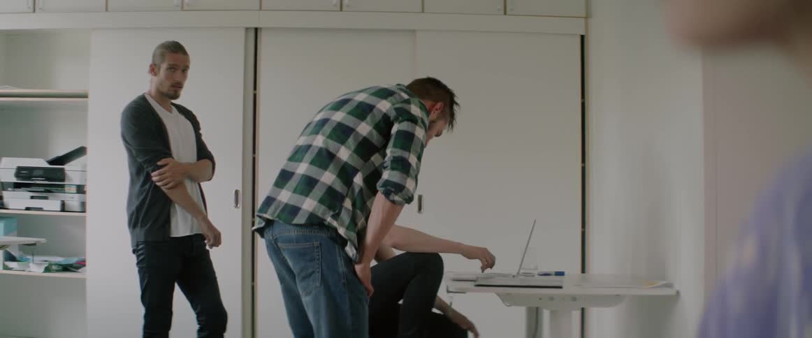 A man sits on a chair looking at a laptop as two other men are nearby
