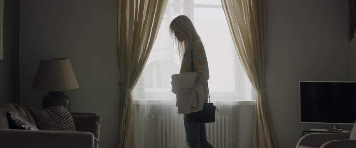 A young blonde woman walks slowly near a window inside a living room.