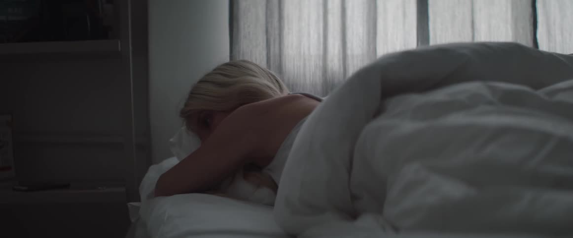 A tired blonde woman sleeps on her side in bed under white sheets.