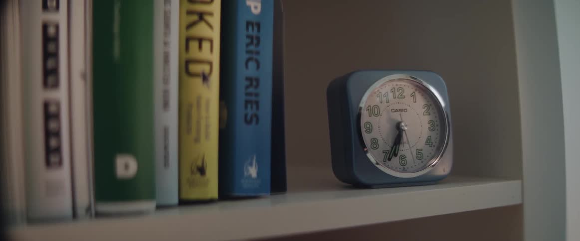 A blue analog alarm clock sits next to books on a white shelf.