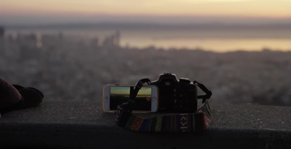 A camera and an iphone sit on a ledge overlooking a city by the bay