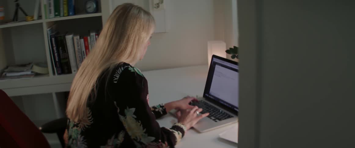 A blonde woman sits at home desk typing on laptop at night.