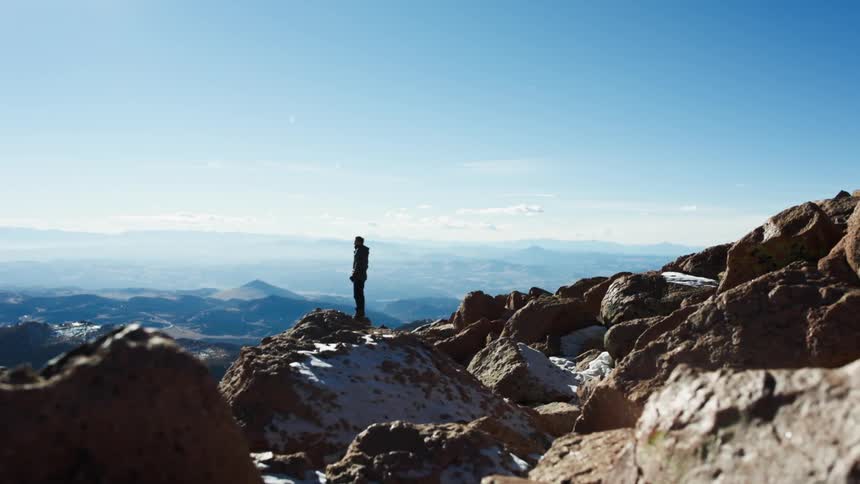 A man stands on rocks on top of a mountain and looks ahead