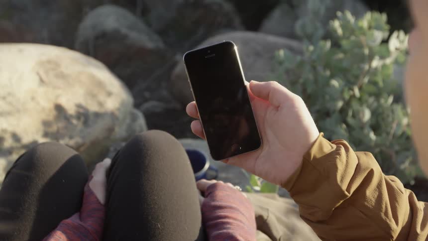 A man staring at the blank screen on his phone as he sits with a woman on top of small boulders