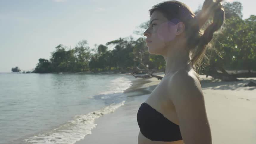 A woman is twisting her hair up into a knot as she stands on a beach during the day
