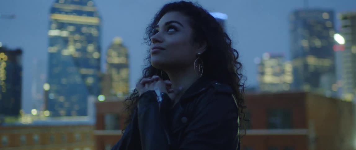 A woman clasps her hands and looks up at the city sky