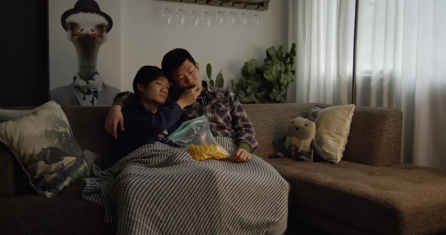 An asian couple are sitting on a couch watching tv and eating snacks