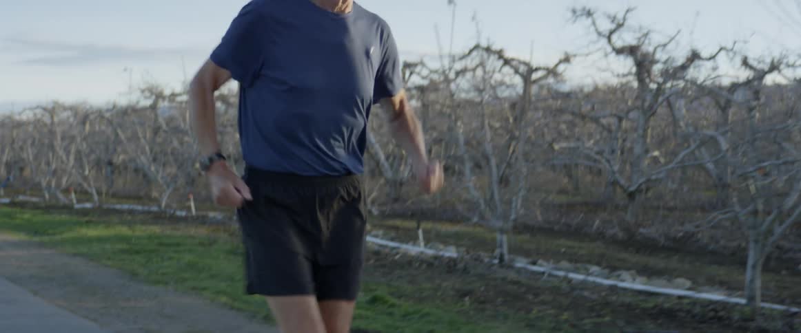 Elderly man jogs down a paved road surrounded by trees