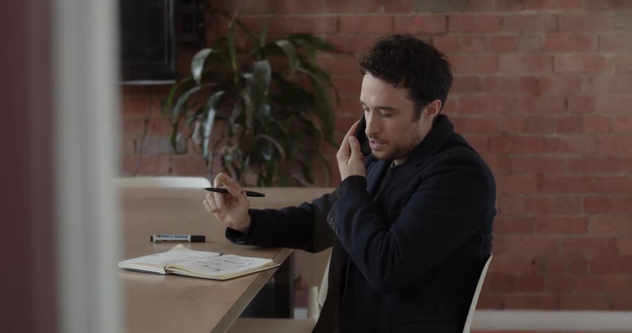 A man is sitting at a desk with a notepad and pen talking on a phone in daylight