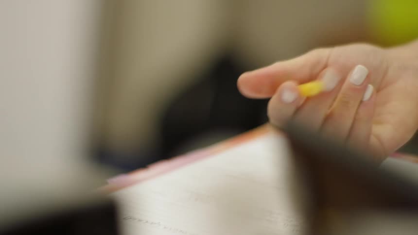 A woman is clicking a pen closed as she writes in a notebook at a desk in daylight