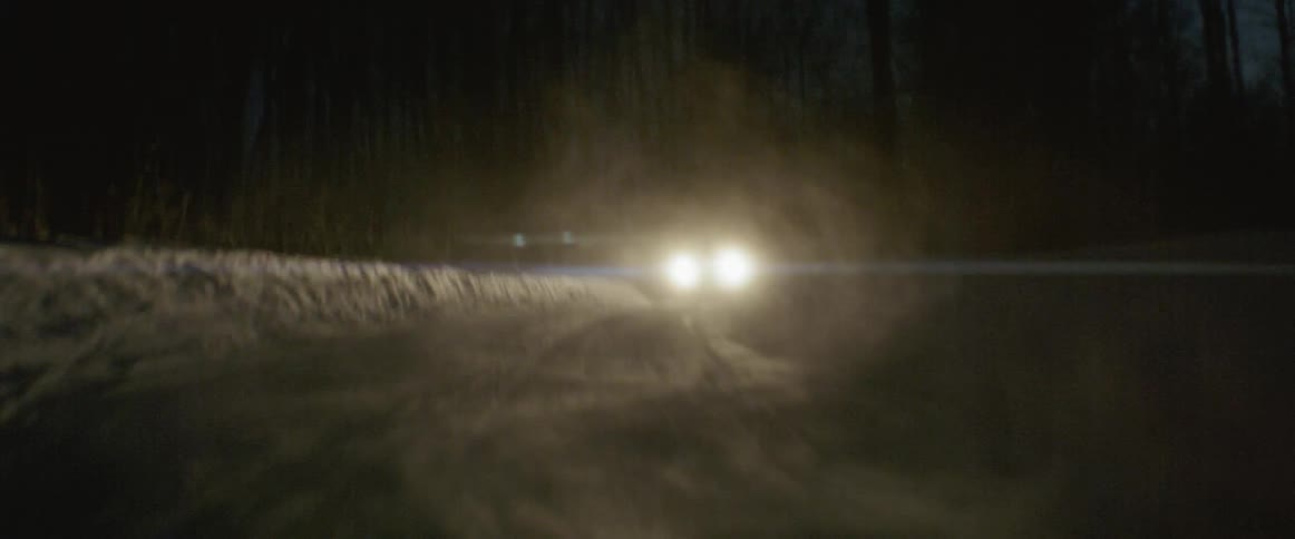 A vehicle is kicking up snow behind it as it drives down a road at night in winter