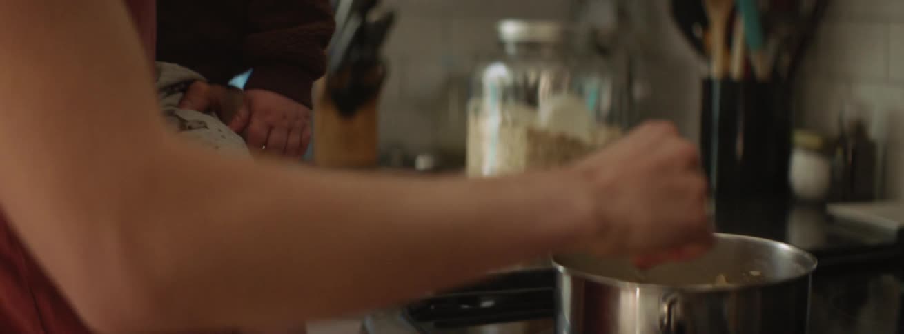 A woman holding a baby prepares food in a pot