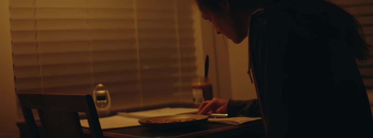 A woman sits at a kitchen table at night writing on a paper and eating