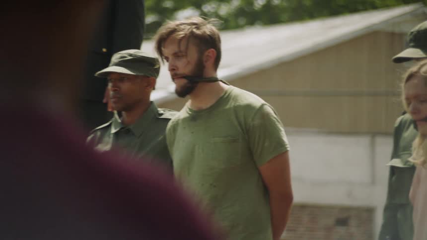 A bound and gagged man and woman stand outside with military people