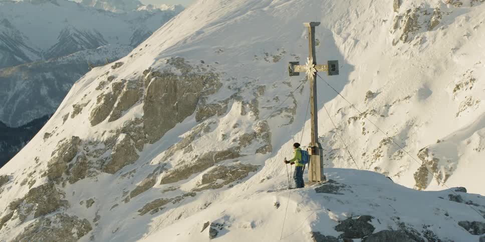 Man holds ski poles as he stands next to a cross on a snowy mountain