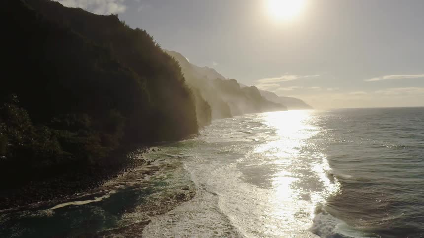 The sun is shining down on waves as they lap the base of cliffs