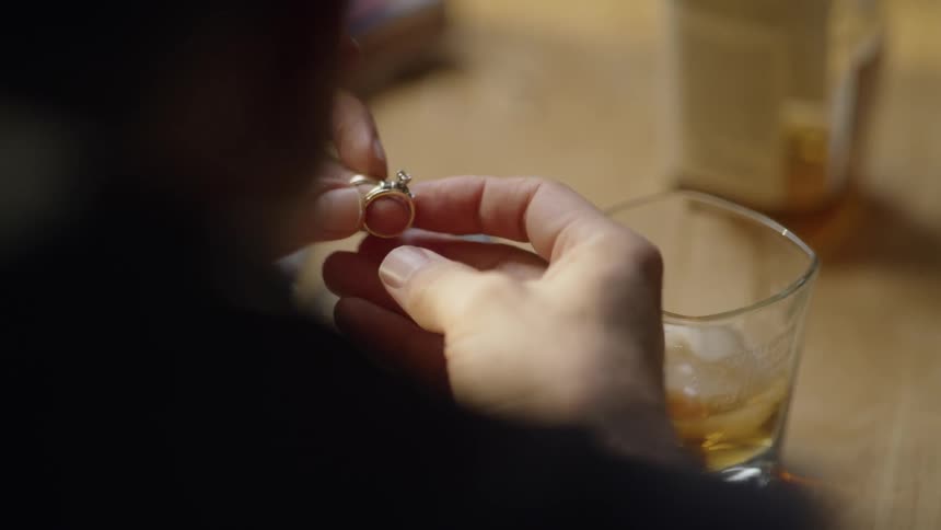 Man sits at table drinking whiskey and looking at wedding rings