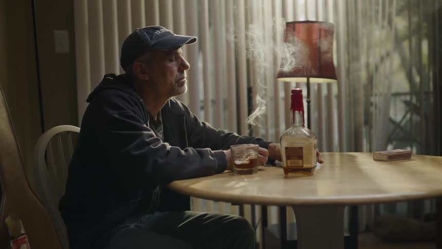 A man sitting at a table with a liquor bottle and glass blows out a candle