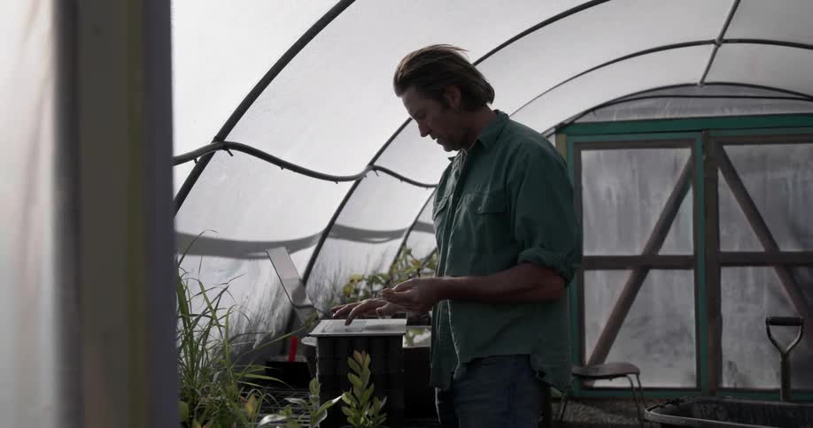 A man in a greenhouse picks up a plant and types info into a laptop