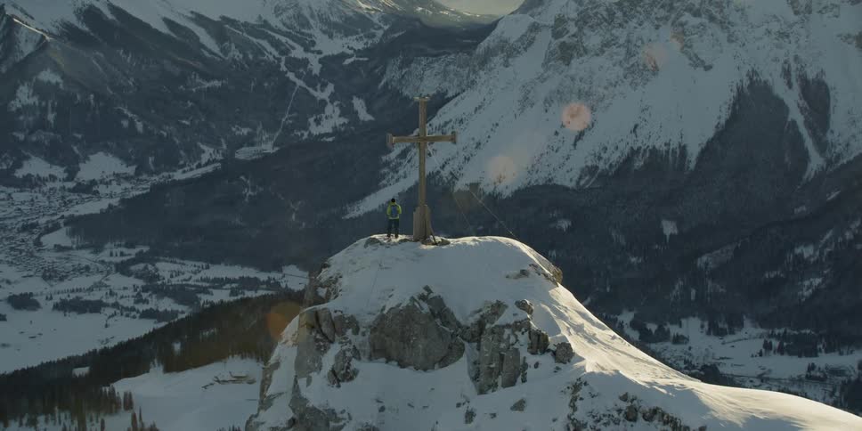 A hiker stands next to a cross on a snowy mountain peak