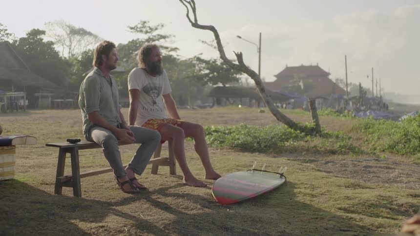 Two men sit on a bench next to a surfboard