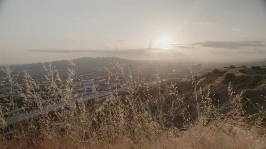 The sun sets over a city and grassy hills