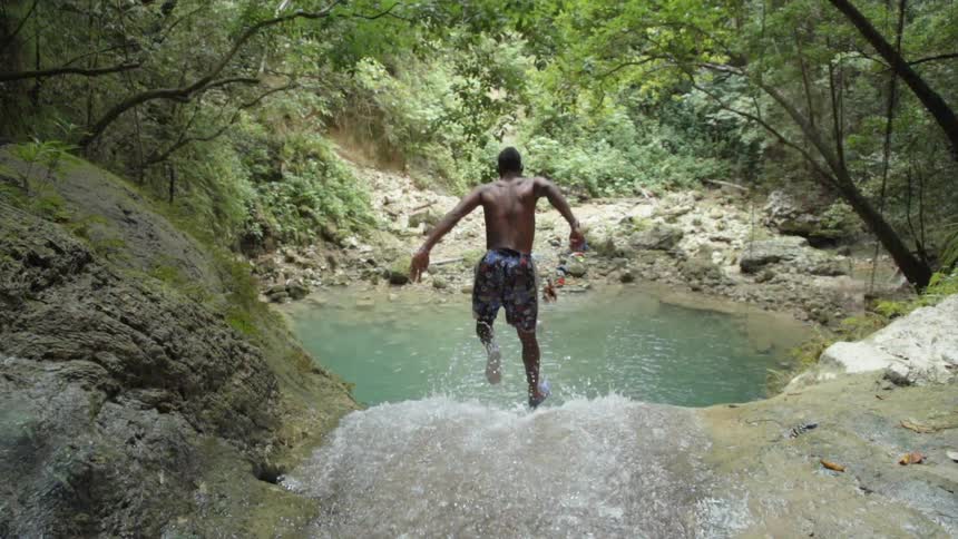 A shirtless man in shorts jumps off a waterfall